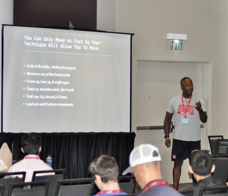 Lawrence Superstar Terry teaching track-and-field technique during a Newberry College coaching presentation with an audience and projected instructional slide.